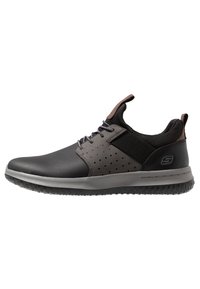 Zapatilla negra y gris con secciones perforadas, panel lateral elástico, suela de goma texturizada, puntera redonda y acentos marrones contrastantes.