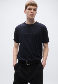 Tengerészkék pamutpóló rövid ujjakkal, enyhe fekete "BOSS" logóval az elején. A modell fekete nadrággal és övvel viseli.