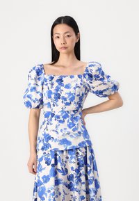 Blouse florale en tissu léger, avec des fleurs bleues sur un fond clair, décolleté carré et manches bouffantes.