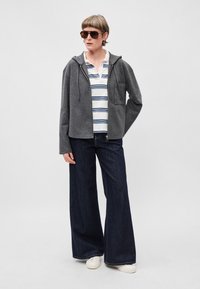 Šedá mikina na zips, biela polo košeľa s modrými pruhmi, tmavo široké džínsy a biele tenisky. Model s okuliarmi stojí pred jednoduchým pozadím.