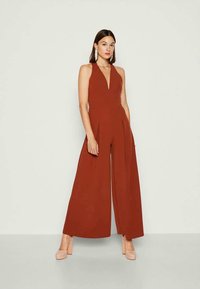 Jumpsuit v rezavé barvě s hlubokým výstřihem do V, širokými nohavicemi a záhyby. Vyroben z hladkého materiálu, doplněný o světle béžové platformové podpatky.