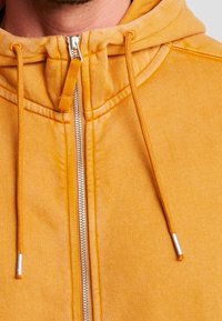 Sudadera con capucha amarilla que presenta una cremallera metálica, cordones ajustables y detalles de costura visibles en un tejido suave.