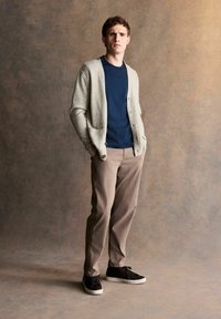 Light gray cardigan, navy tee, tan pants, and black sneakers. Cardigan features buttons; fabric appears soft. Standing pose against textured background.