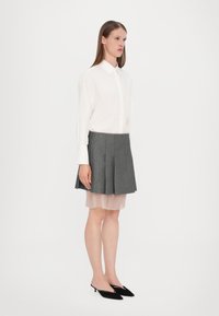 Chemise blanche à col, jupe plissée grise et chaussures à talons noirs. Jupe superposée sur une sous-couche plissée rose clair. Textures lisses.