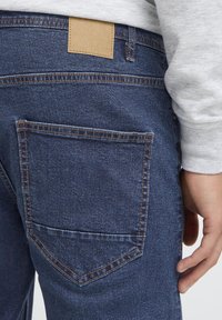 Tamno plave traperice s džepom na stražnjoj strani s kontrastnim šavovima i smeđim kožnim detaljem na struku. Glatka tekstura, uski kroj.