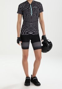 Jersey de ciclismo negro con patrones geométricos, de manga corta, con cremallera; combinado con pantalones cortos negros que tienen una banda texturizada de cuadros. Casco negro en la mano.