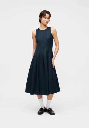 Ung kvinde stående med hænderne i lommerne iført en ærmeløs mørkeblå midikjole, hvide sokker og sorte loafers mod en ensfarvet baggrund.