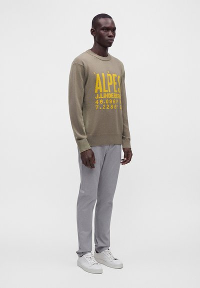 Homme portant un pull beige avec un texte jaune, un pantalon gris et des baskets blanches, debout devant un fond blanc uni.