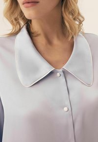 Lawendowa satynowa bluzka z dużym okrągłym kołnierzem z białym wykończeniem i zapięciem na guziki przy szyi. Gładka tekstura o delikatnym połysku.