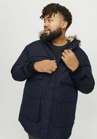 Marine parka met een bontgevouwde capuchon, vooraan met rits en drukknopen. Bevat twee borstzakken en twee zijzakken, evenals geribde manchetten.