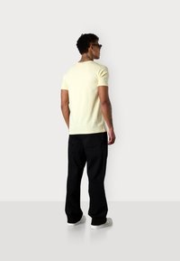 T-shirt ajusté jaune clair à manches courtes, associé à un pantalon noir. Le modèle porte des baskets blanches et des lunettes de soleil noires, affichant un tatouage sur le cou.