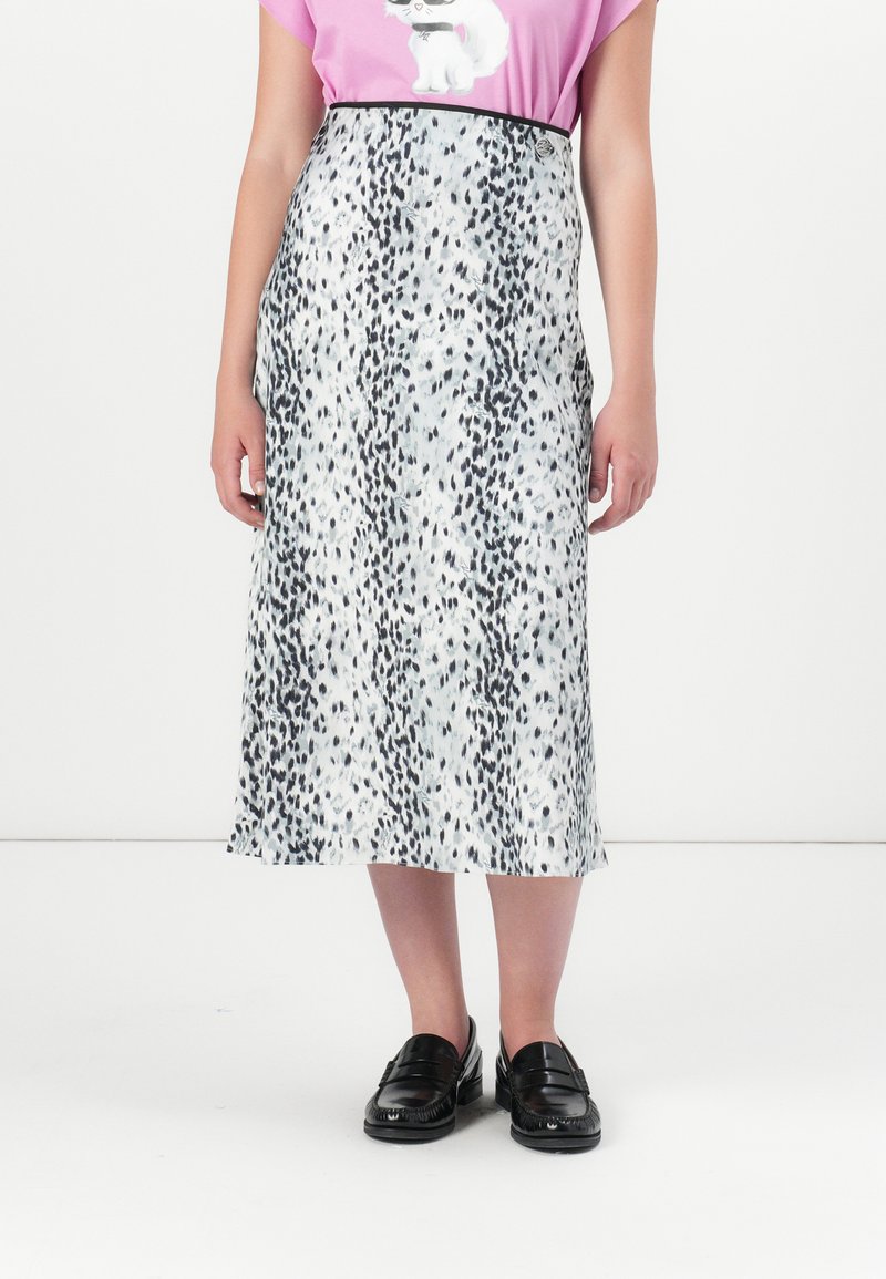 Une personne se tient debout portant un t-shirt rose avec un motif de chat et une longue jupe à pois noirs, accompagnée de mocassins noirs.