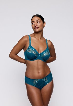 Vrouw draagt tealkleurig lingerie met bloemenborduurwerk, staand tegen een effen lichtgrijze achtergrond, kijkt naar links met haar handen achter haar rug.