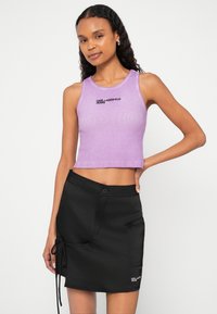 Vijoličasta skrajšana majica z rebrasto teksturo in črnim vezenim logotipom, kombinirana s črno krilo s pojavnim gumbom in stranskim žepom.