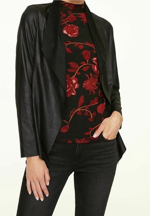 Kvinde iført sort draperet jakke med åben front over sort top med rødt blomsterprint og sorte jeans, den ene hånd i lommen.