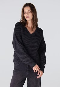 Zwarte, getextureerde gebreide trui met een V-hals en lange, volumineuze mouwen. Geribde zoom en manchetten voegen detail toe. Draagt men over donkere jeans.