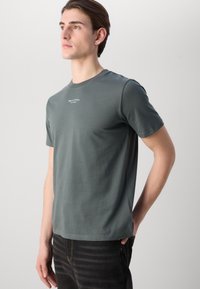 Mørkegrønn bomulls T-skjorte med rund hals og korte ermer, med en liten hvit logo på brystet. Brukt med svarte jeans.