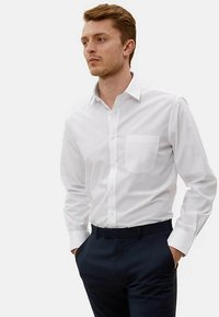 Bílá košile s dlouhým rukávem, s kapsou, límcem na knoflíky a rovnými lemy. Tkanina působí hladce, doplněná tmavými kalhotami.