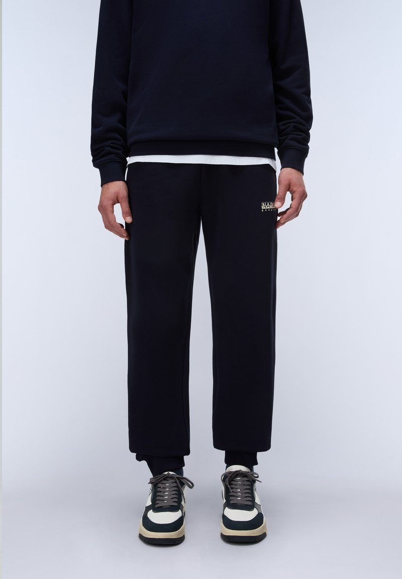 Personne portant un pantalon de survêtement noir court, un sweat noir sur une chemise blanche et des baskets noires et blanches, debout sur un fond clair uni.