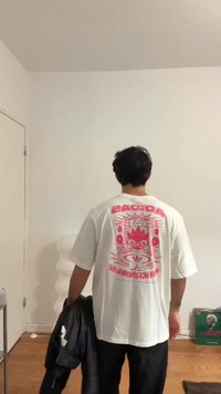 Balti marškinėliai su ryškiomis rožinėmis grafikos detalėmis nugaroje. Laidi forma su trumpomis rankovėmis. Juoda striukė laikoma kairėje rankoje.