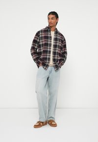 Karirana flanelna srajca v temnih tonih, oversized kroja, v kombinaciji s svetlo modrimi kavbojkami in rjavimi supergami, kar ustvarja sproščen, vsakdanji stil.
