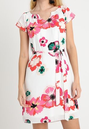 Vrouw draagt een witte jurk met korte mouwen, bedrukt met rode, roze en groene bloemen, en een ceintuur in de taille, staand tegen een effen achtergrond.