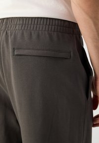 Close-up van een persoon in donkergrijze elastische broek met een horizontale achterzak en een wit shirt erin gestopt.