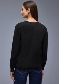 Mujer con cabello largo y castaño, que lleva un suéter negro de manga larga y unos vaqueros azules, vista de espaldas contra un fondo gris.