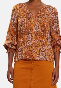 Blouse ample à col V en jaune moutarde avec des motifs floraux, des manches bouffantes et des détails plissés. Associée à une jupe orange assortie.