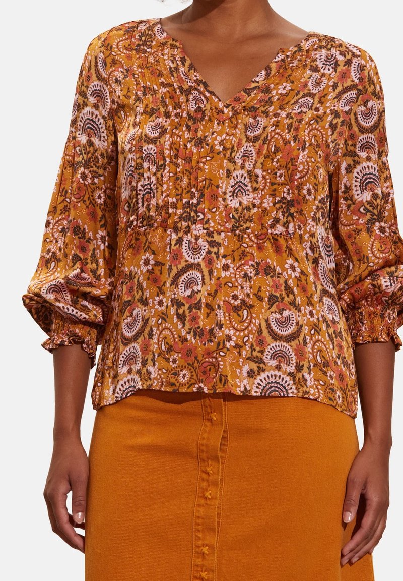 Blouse ample à col V en jaune moutarde avec des motifs floraux, des manches bouffantes et des détails plissés. Associée à une jupe orange assortie.
