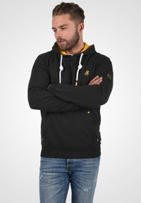 Zwarte hoodie met gele accenten, witte trekkoorden en een voorzak. Bevat een geborduurd logo op de borst en de rechter mouw.