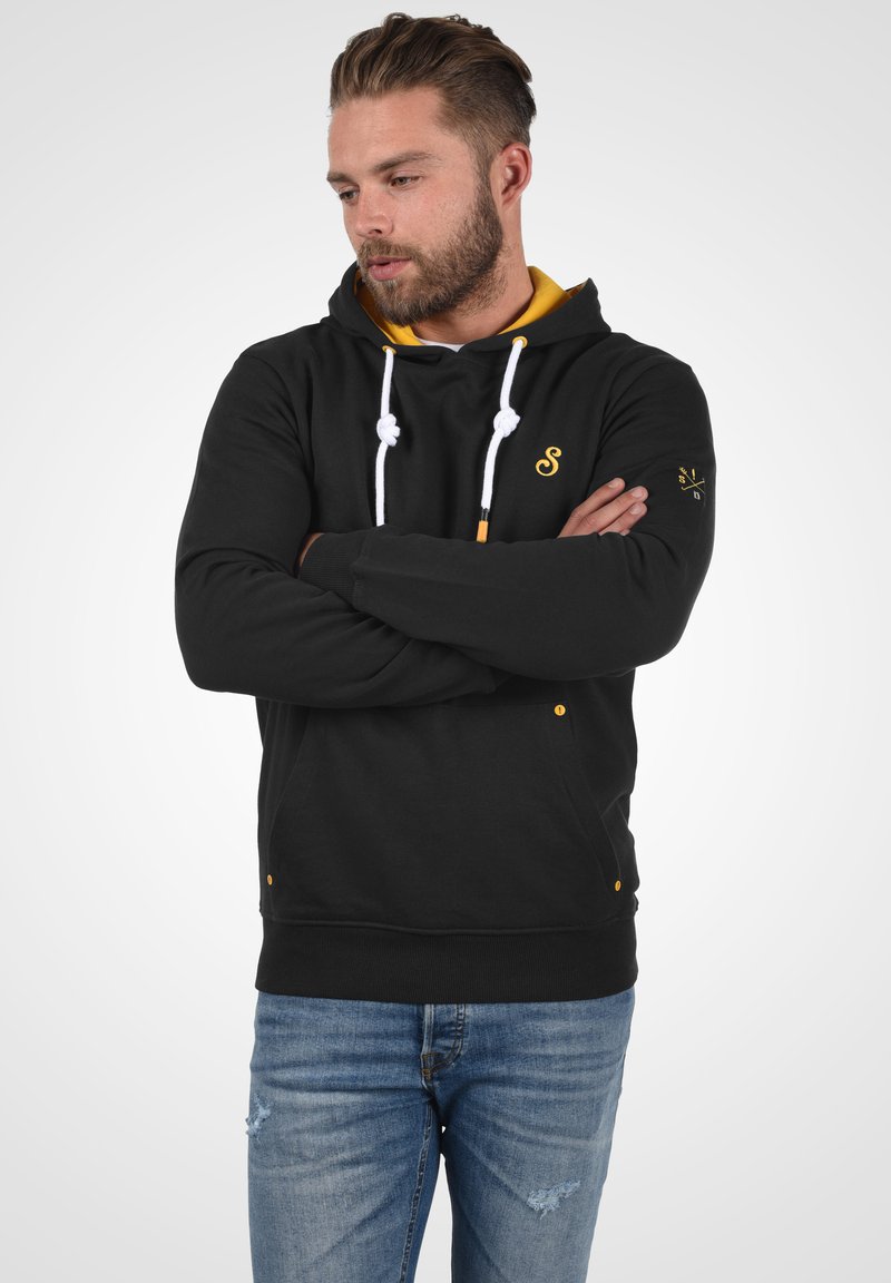 Zwarte hoodie met gele accenten, witte trekkoorden en een voorzak. Bevat een geborduurd logo op de borst en de rechter mouw.