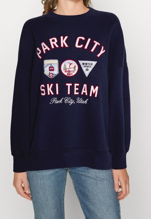 Granatowa bluza z napisem „Park City Ski Team” i naszywkami związanymi z narciarstwem, noszona z jasnoniebieskimi dżinsami na stojącej osobie.