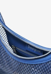 Blaue Tasche mit einer äußeren Schicht aus Netzstoff und Lederfutter, ausgestattet mit silbernen Reißverschluss-Schließen und einem Logoschild auf der Innenseite. Sichtbares, strukturiertes Muster.