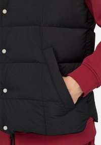 Juodas pūkinis liemenė su lygiu paviršiumi, sidabro spalvos mygtukai ir šoninės kišenės, dėvimas ant bordo spalvos ilgomis rankovėmis marškinėlių.