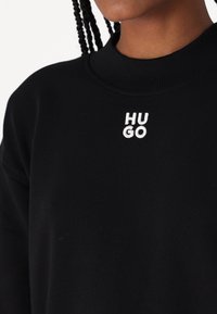 Černá mikina s ribbeným límcem a bílým vyšitým logem "HUGO" na přední straně. Materiál vypadá měkce a má hladkou texturu.
