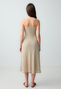 Knielange, aansluitende beige jurk met een verfijnd breipatroon, dunne spaghettibandjes en een gladde textuur. Minimalistisch ontwerp, geen zichtbare sluitingen.
