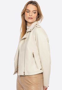 Chaqueta de motorista de imitación de cuero color beige claro con detalles en oro, cuello a presión y forma ajustada. Textura suave y bolsillos laterales con cremallera.