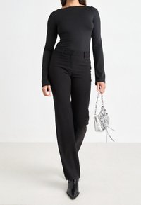 Schwarzes langärmeliges Oberteil mit einem Rundhalsausschnitt, kombiniert mit maßgeschneiderten schwarzen Hosen. Als Accessoires dienen eine kleine metallisch silberne Handtasche und spitze Stiefel.