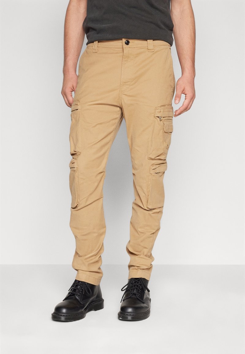 Pantalon cargo beige avec plusieurs poches latérales, coupe droite, en tissu de coton résistant, associé à des bottes à lacets noires.