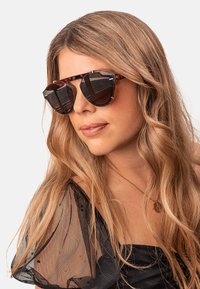 Tortoiseshell sunglasses with a bold, angular design, dark lenses, and gold-tone accents. Accessorized with a sheer black top.