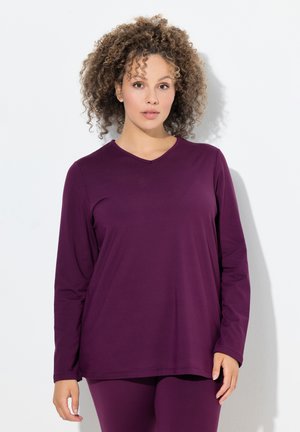 Kvinna med lockigt hår som bär en långärmad lila V-ringad tröja och matchande byxor, stående mot en vit bakgrund.