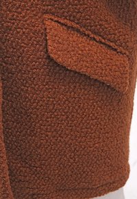 Brunt texturerat tyg med en mjuk, luddfinish. Har en lappficka på vänster sida, formad för att smälta in i den övergripande designen.