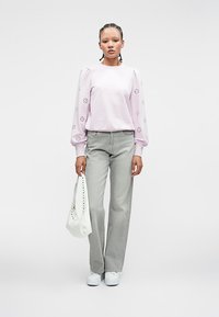 Vrouw staat met een licht paarse lange mouwen top met bloemen uitsneden, grijze wijde jeans, witte sneakers en houdt een witte handtas vast.