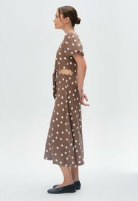 Braunes Punktekleid mit Puffärmeln und seitlichem Ausschnitt, mit einem fließenden Rock und einer Bindung in der Taille, kombiniert mit schwarzen Ballerinas.
