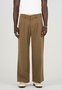 Pantalones plisados marrones hechos de tela suave, con una cintura alta, piernas anchas y cierre de botón. Combinados con zapatos color crema.