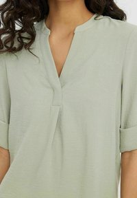 Femme portant un chemisier vert clair à col en V et manches courtes retroussées, dévoilant le haut de la poitrine et des cheveux bouclés foncés.