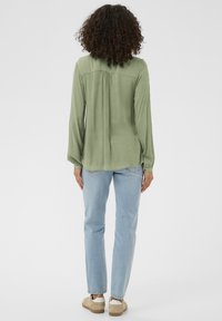 Blouse à manches longues vert clair avec une coupe décontractée, dotée d'un pli au dos, associée à un jean droit bleu clair et des chaussures beige.