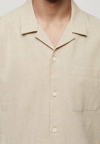 Beige linnen overhemd met korte mouwen, voorzien van een uitgesneden kraag, vijf knopen aan de voorkant en een borstzak aan de linkerkant. Gestructureerde stof.