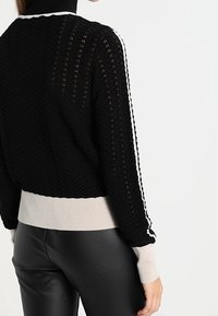 Schwarzer Strickpullover mit Fischgrätenmuster, weißen Akzentstreifen an den Ärmeln und einem beigen, gerippten Saum.